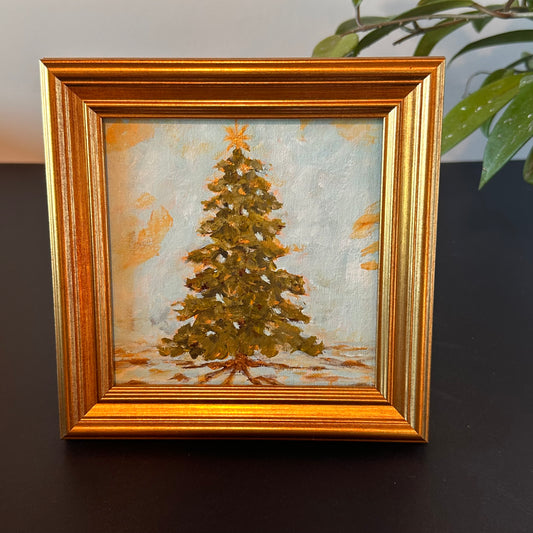 Framed painting of a Christmas tree on a dark surface with a plant in the background